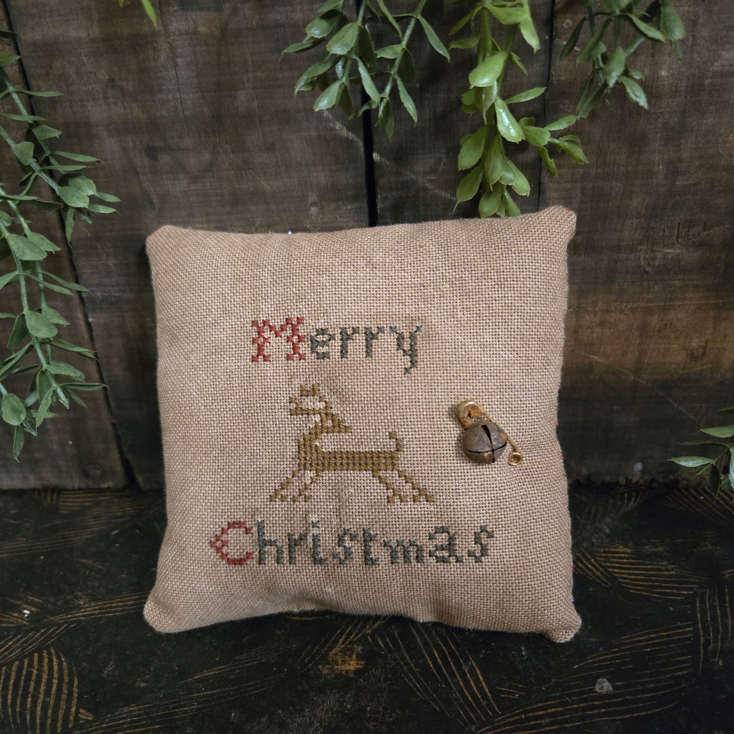 Decorative pillow with cross-stitched text on a wooden surface with greenery