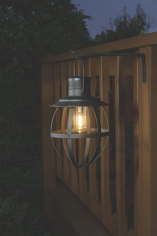 A metal hanging lantern with a cylindrical shape and a loop at the top, featuring a lit candle inside, mounted on a wooden fence post.