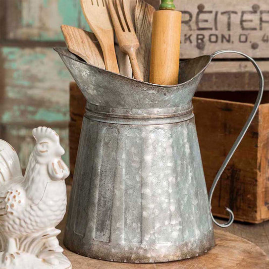 A metal milk pitcher with a handle, filled with utensils and dried flowers, placed on a wooden surface with a chicken figurine and a sign in the background.