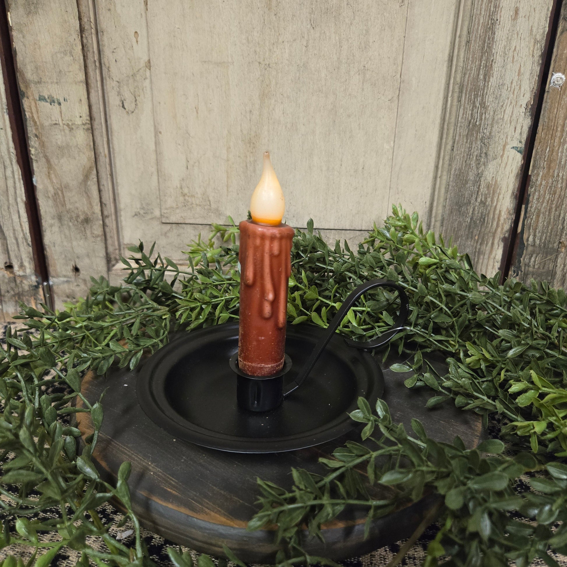 A metal taper candle holder with a lit candle surrounded by green foliage on a round table.