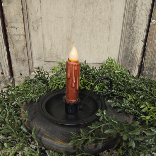 A metal taper candle holder with a lit candle surrounded by green foliage on a round table.