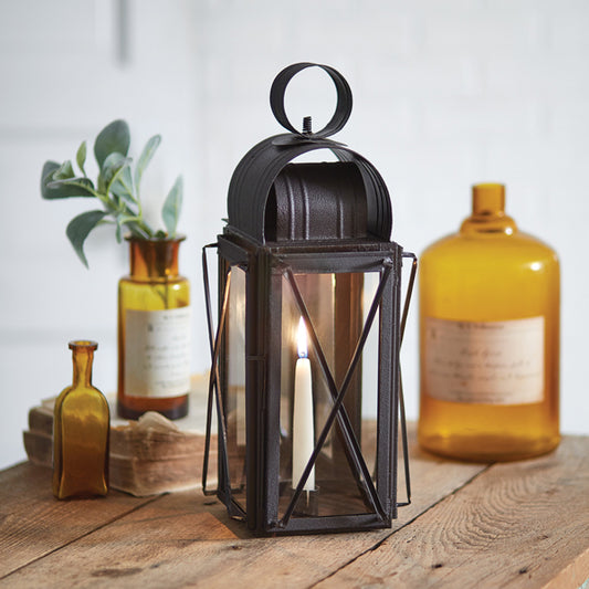 A black metal milkhouse lantern with a mirrored back and glass panels on three sides, featuring a lit candle inside, placed on a wooden surface with various decorative items in the background.
