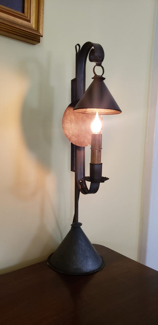 A Mini Bell Tin Table Light with a reflector, featuring a bronze-colored base and a dark-colored bell-shaped shade, placed on a wooden surface against a wall.