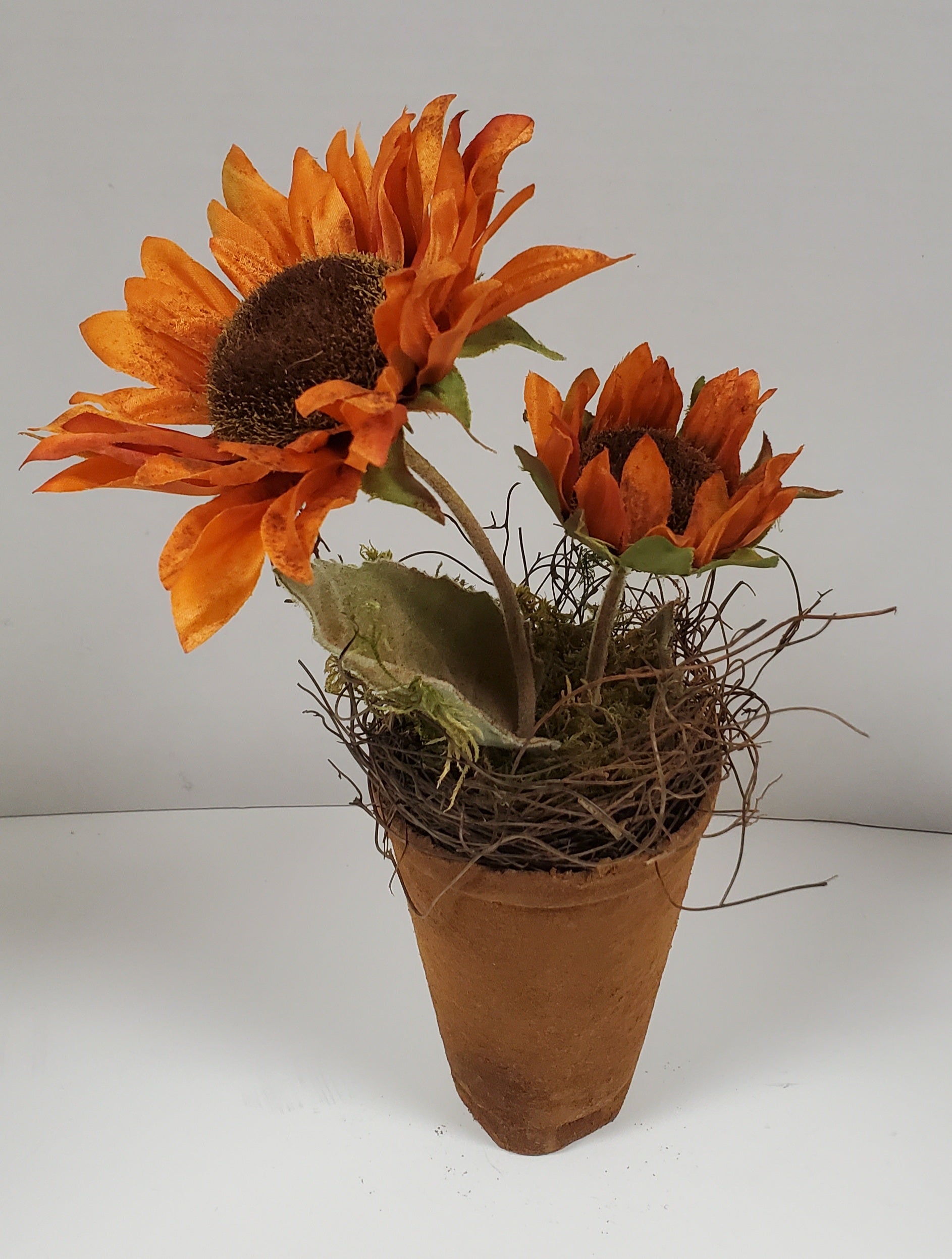 A brown pot with cinnamon coating and wax dipped orange sunflowers with brown centers and green leaves.