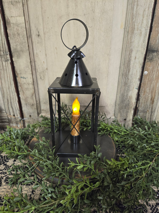 A black mini tin lantern with cross wires, displaying a lit candle inside, set on a bed of greenery.