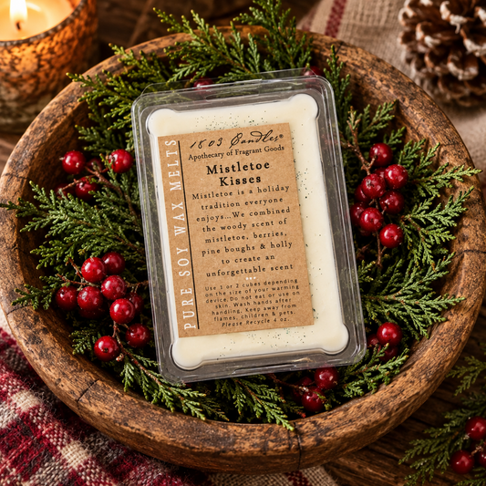 A square clear plastic clamshell wax melt package labeled 'Mistletoe Kisses' by 1863 InnLabs Apothecary of Fragrance Candles, placed on a rustic wooden tray surrounded by green pine branches and red berries.