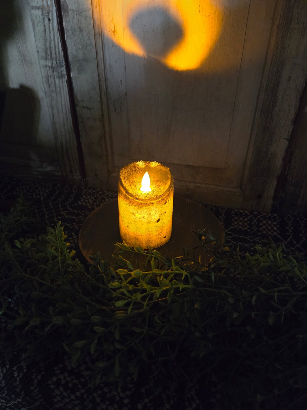 Candle on wood riser with greenery in a dimly lit room.