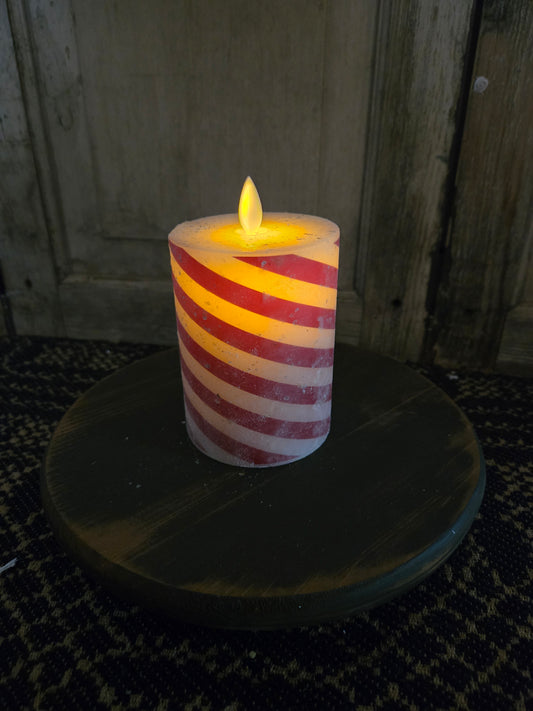 A battery-operated candle with a moving flame effect, designed to resemble a candy cane, placed on a round wooden pillar.