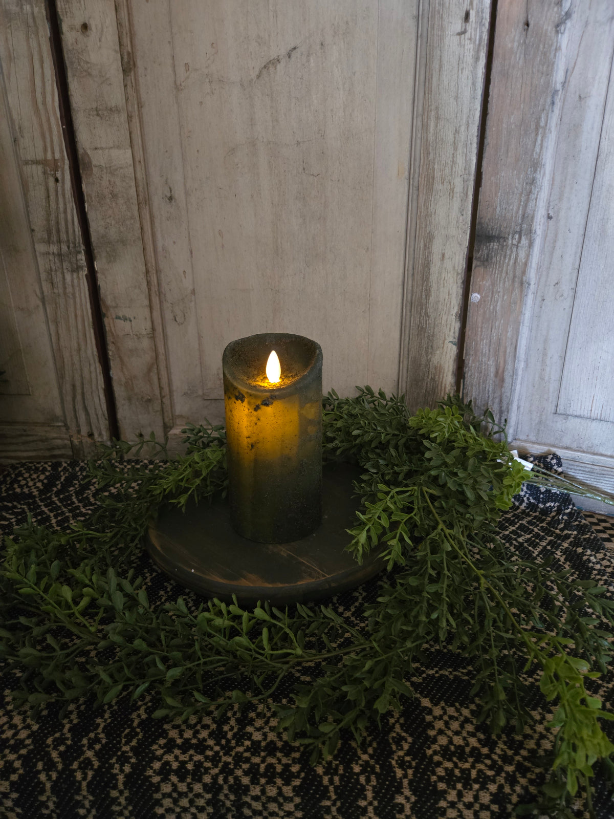 Candle on wood riser with greenery in front of a weathered wood background.