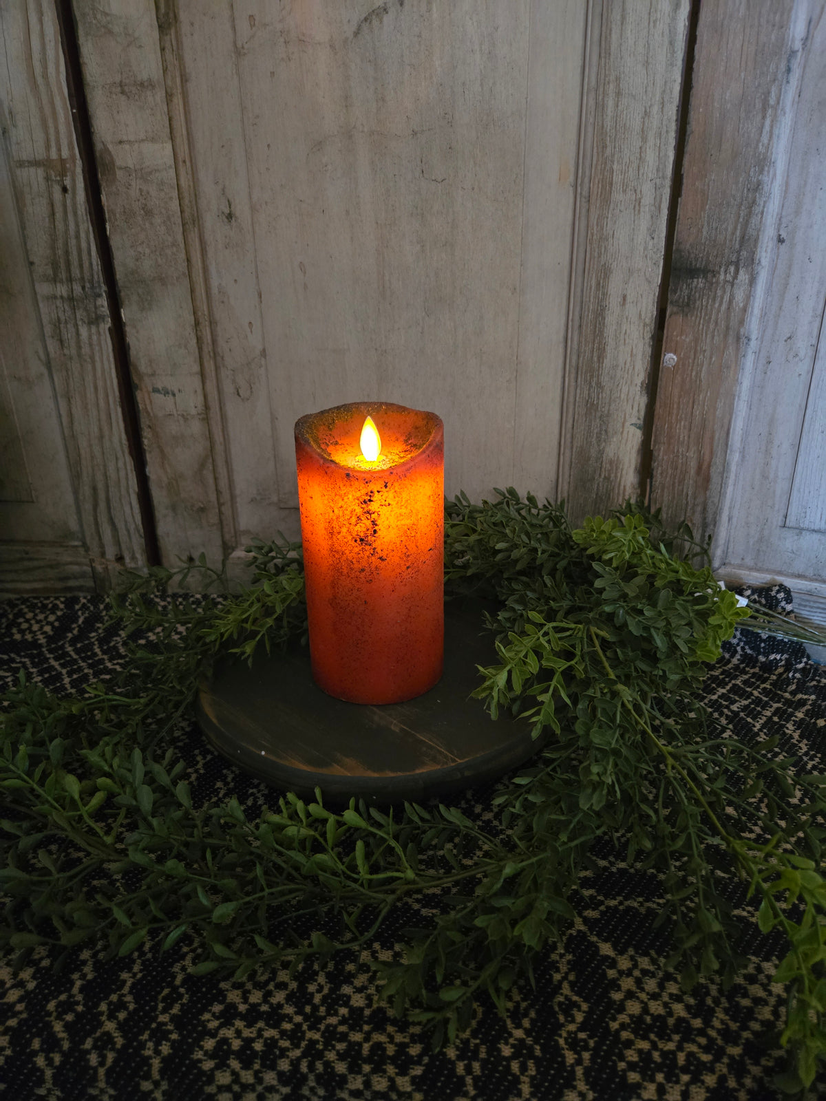 Pumpkin colored candle on wood riser with greenery in front of a weathered wood background.