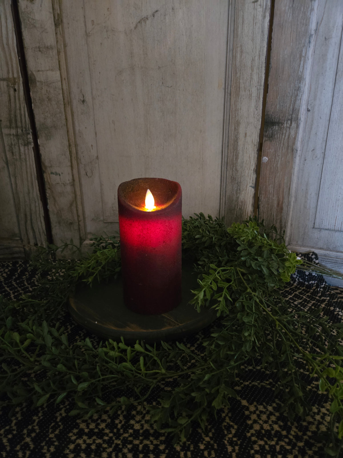 Red candle on wood riser with greenery in front of a weathered wood background.