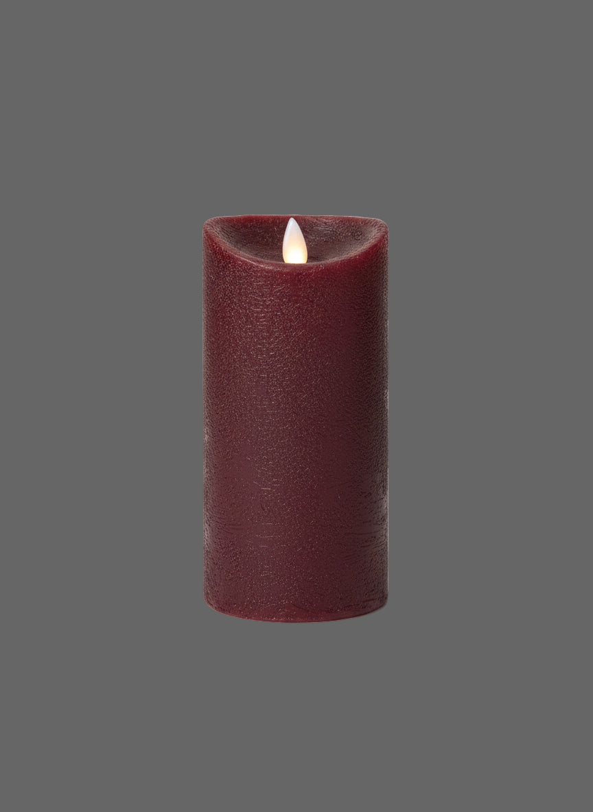 A red pillar candle with a moving flame, surrounded by green foliage, placed on a dark textured surface against a wooden background.