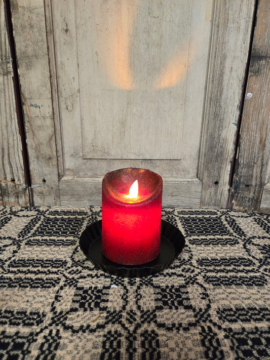 A red pillar candle with a moving flame, placed on a textured black mat, with an open door in the background.