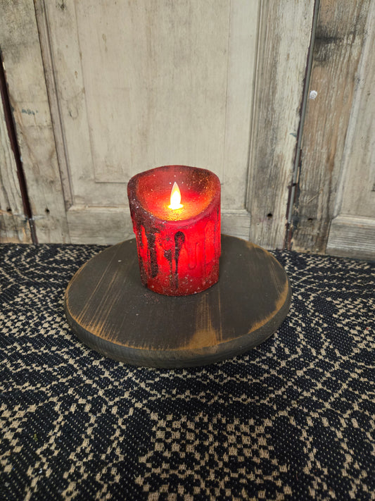 A red mica pillar candle with an illuminated flame, placed on a round, dark wicker mat, with a rustic wooden background and a door in the background.