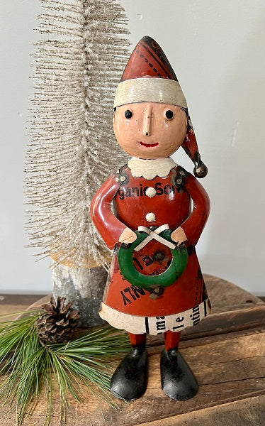 A figurine of Mrs. Claus holding a wreath, wearing a red and white outfit with a Santa hat, placed on a wooden surface with pine cones and a white decorative item in the background.