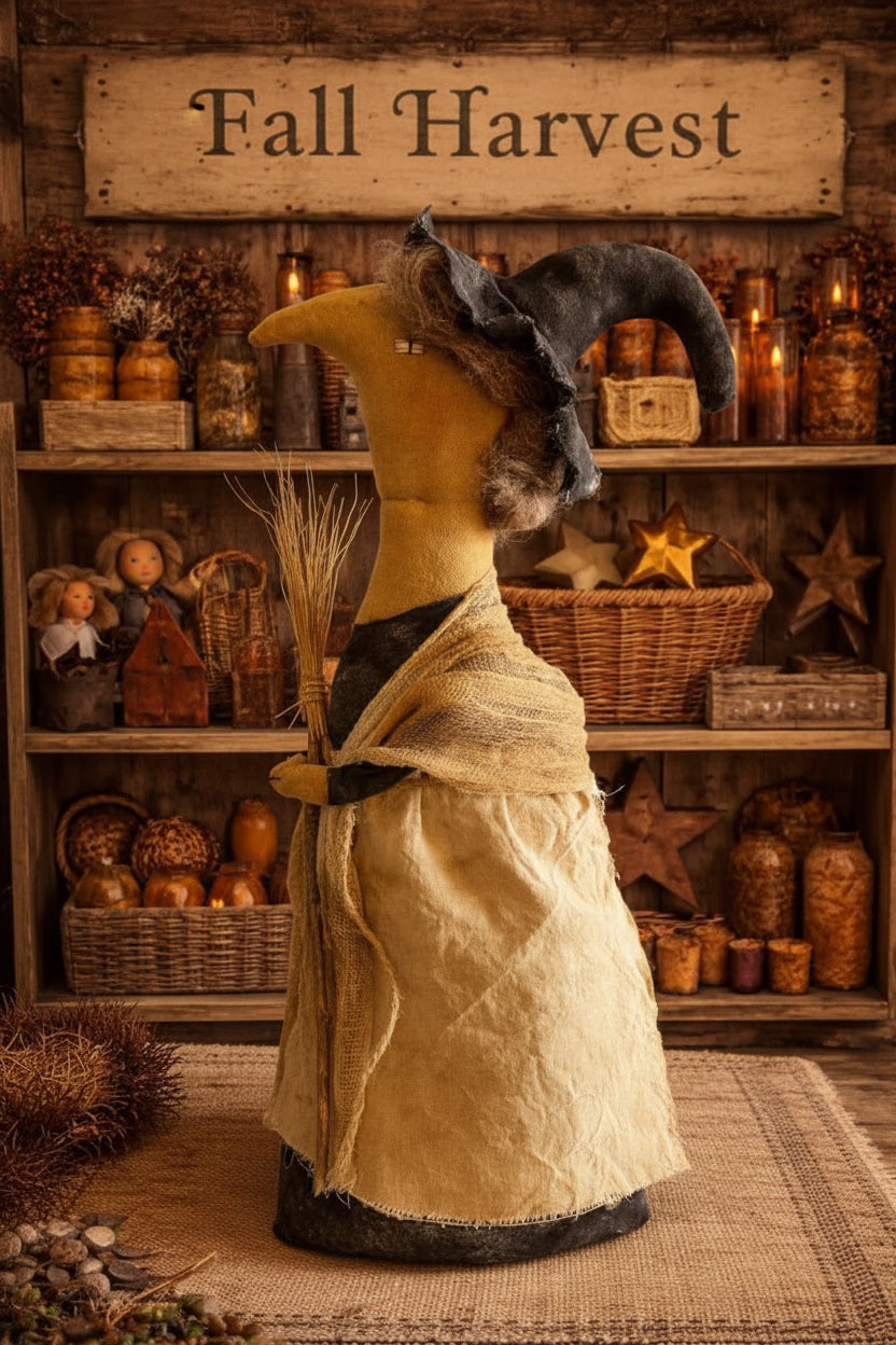 A rustic witch figurine made from natural materials, featuring a yellow body, black hat, and beige dress, standing in front of a rustic wooden shelf display.