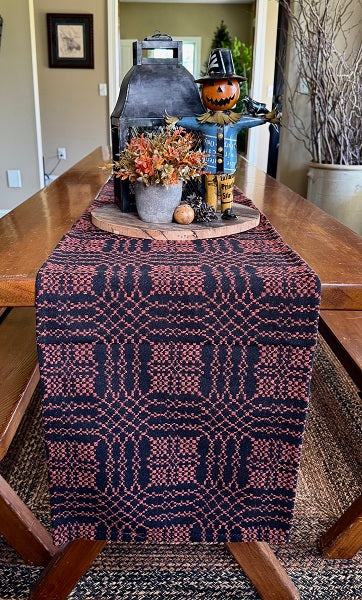 A woven table runner with a dark navy and dusty rose geometric pattern draped over a wooden dining table, styled with autumn decorations including a lantern, a small pumpkin, and a vase of fall foliage.