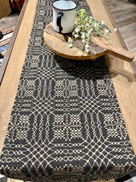 A black and cream woven table runner with a geometric pattern, styled on a wooden dining table with a rustic mug and dried flowers on a wooden board.