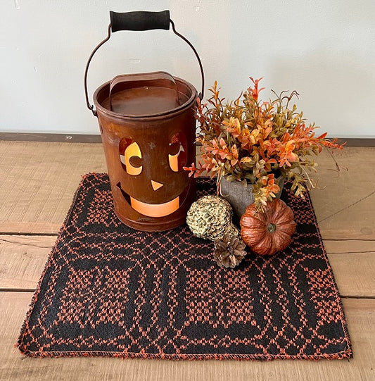 A square placemat with a black and rust color scheme, featuring a jack-o-lantern design, placed on a wooden surface. Autumn-themed decorations, including pumpkins and dry leaves, are placed on top of the placemat.
