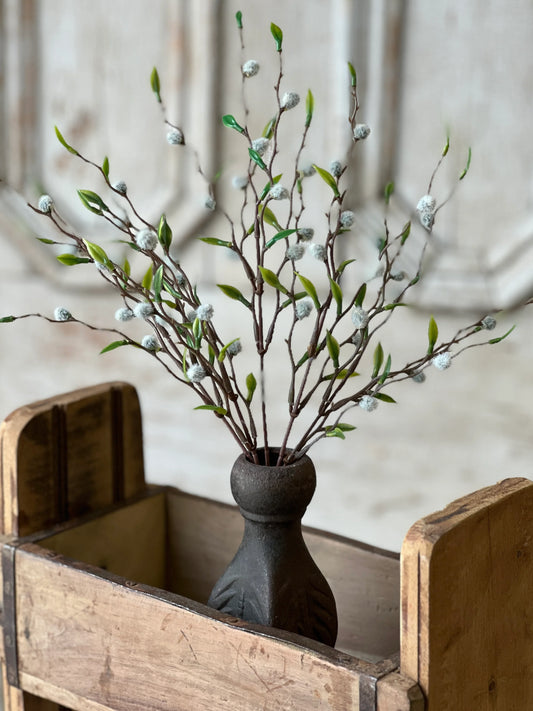 A decorative floral arrangement featuring branches with leaves and cloud-like catkins in a black vase, placed inside a wooden crate.