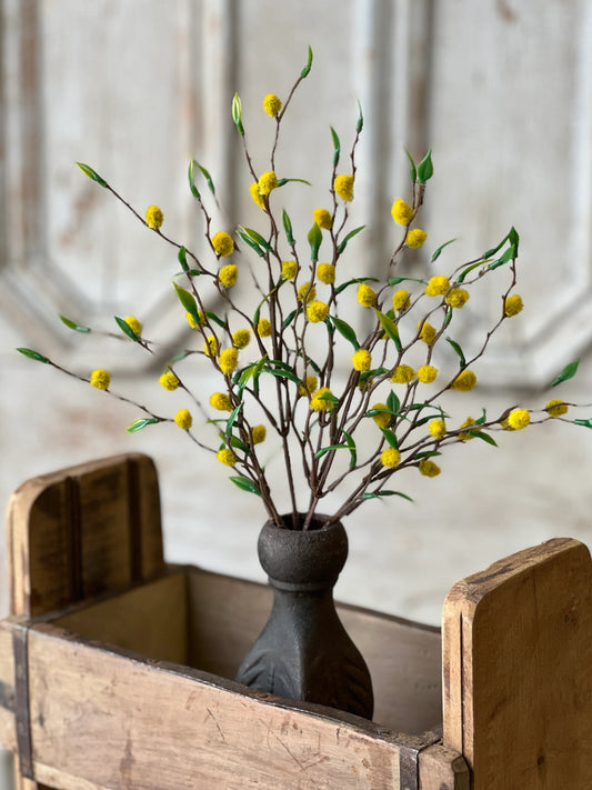 A decorative floral arrangement featuring yellow faux fruits on green stems, placed in a black vase, set against a rustic wooden backdrop.