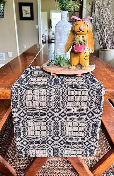 A woven table runner with a black and beige geometric pattern, styled on a wooden dining table with a decorative bunny figurine and a white vase in the background.