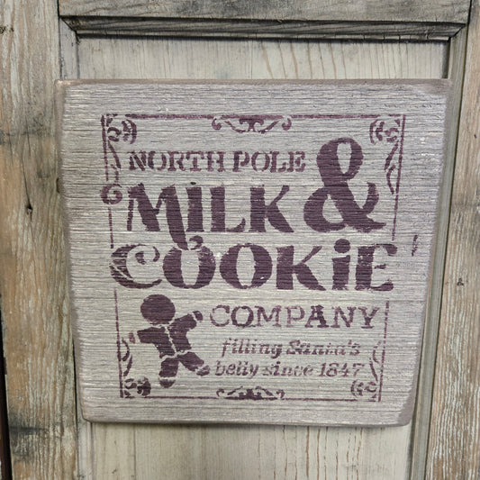 A wooden sign with a vintage design featuring the text 'NORTH POLE MILK & COOKIE COMPANY' and an illustration of a cookie.