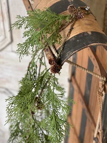 A close-up of a rustic wooden bucket or barrel lid decorated with green evergreen branches, pinecones, and a braided rope, set against a softly blurred indoor background.