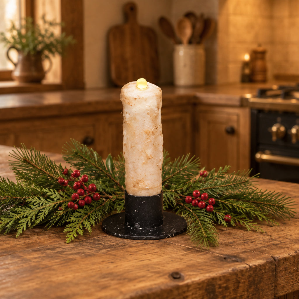 A single faux LED pillar candle with a warm beige wax body and a black base, shown against a background of green pine branches and brown pinecones.