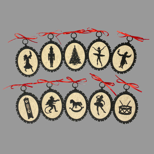 A set of paperboard silhouette ornaments with laser cut designs, featuring characters from The Nutcracker Ballet, such as the nutcracker, mouse king, and Christmas tree, each adorned with a red ribbon and hanging from a red string.
