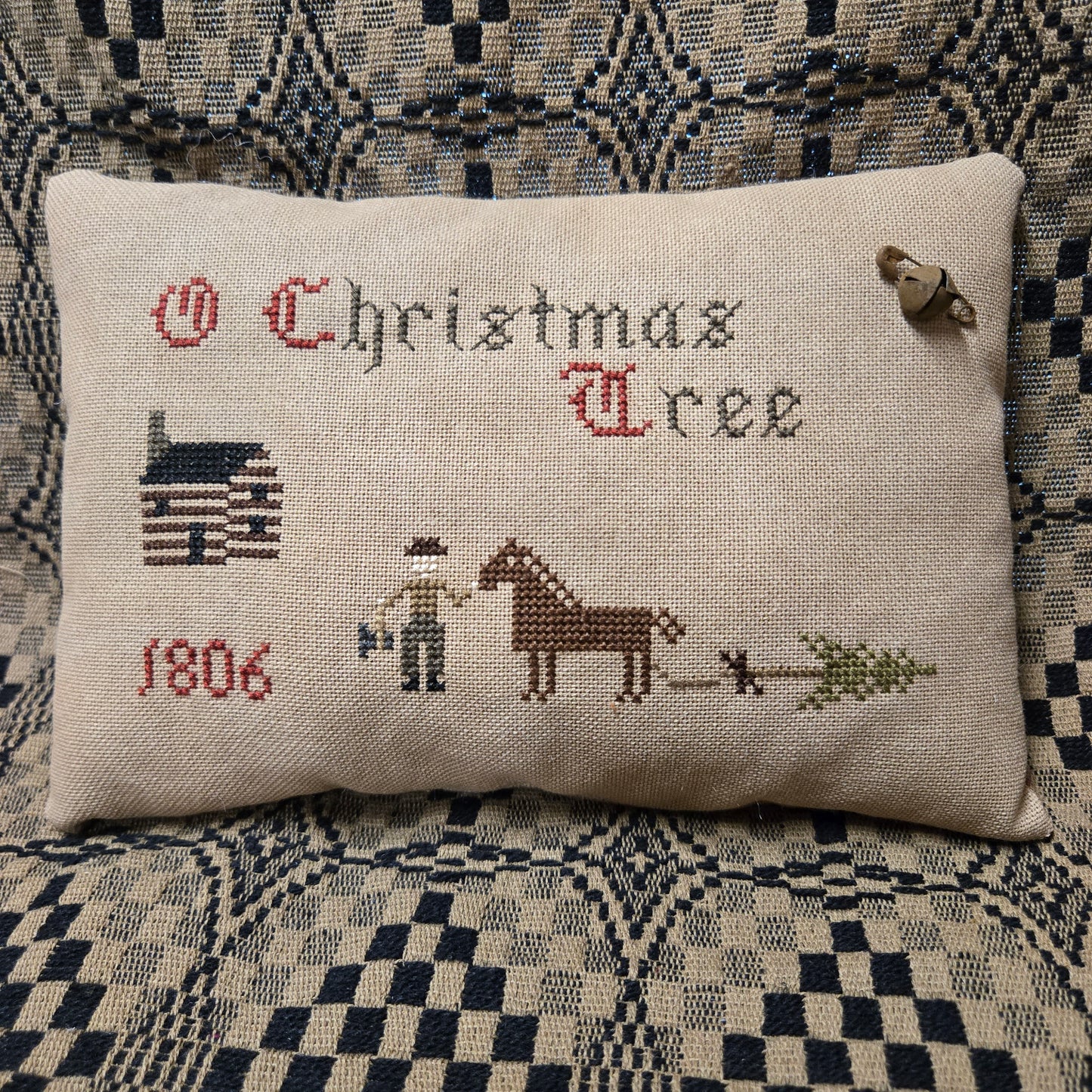 A rectangular cross-stitch pillow on a patterned blue and beige couch. The pillow is made of cream linen fabric and features embroidered text reading 'O Christmas Tree' with a cabin, '1806', a person, a horse, and a Christmas tree.
