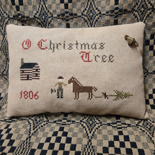 A rectangular cross-stitch pillow on a patterned blue and beige couch. The pillow is made of cream linen fabric and features embroidered text reading 'O Christmas Tree' with a cabin, '1806', a person, a horse, and a Christmas tree.