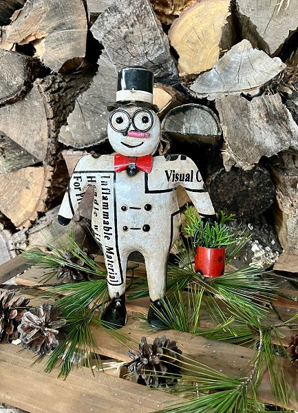A metal snowman decoration with a top hat, bow tie, and the text 'VISUAL' on its chest, surrounded by pine cones and needles on a wooden surface with stones in the background.