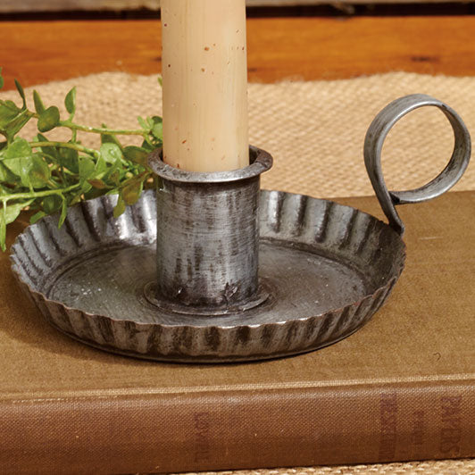 A metal candle holder with an aged finish and crimped edges, featuring a finger loop on the side. The candle is not included.