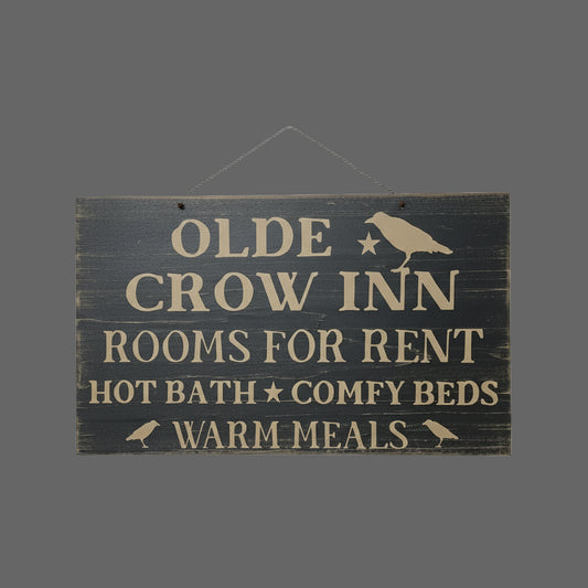 A black wooden sign with mustard lettering that reads 'Olde Crow Inn Rooms for Rent Hot Bath Comfy Beds Warm Meals' with a graphic of a crow perched on a branch.