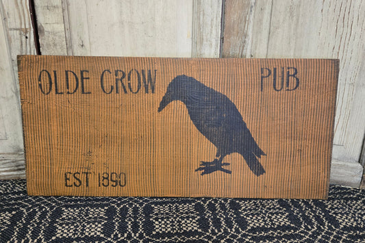 A hand-painted combed sign with 'OLDE CROW PUB' text and an illustration of a crow perched on a fence post, placed against a rustic wooden background.