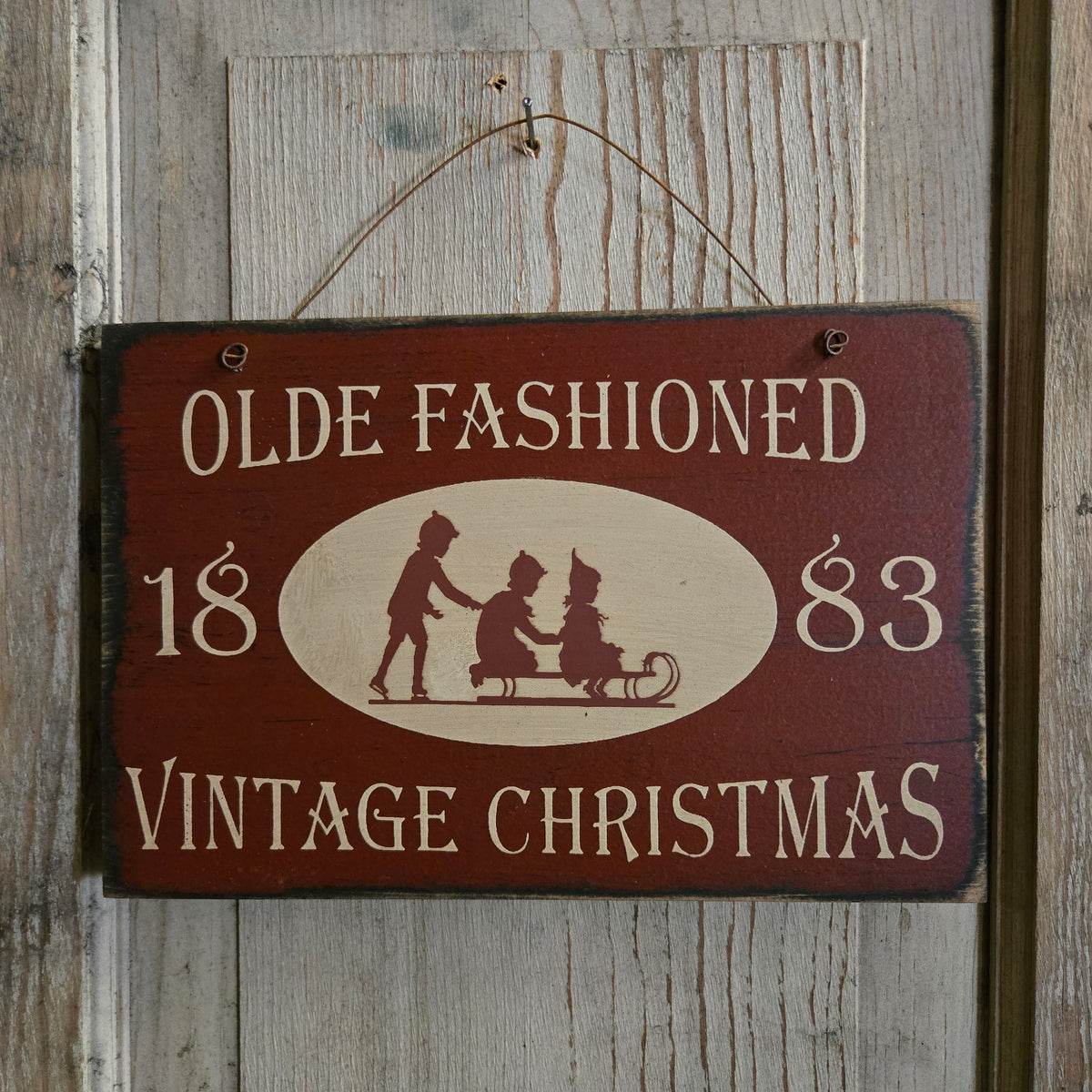 A rustic rectangular wooden hanging sign with a dark red painted background and cream lettering that reads “OLDE FASHIONED 1883 VINTAGE CHRISTMAS,” plus a cream oval with silhouetted figures in a sleigh. The sign hangs against a weathered wood plank wall.