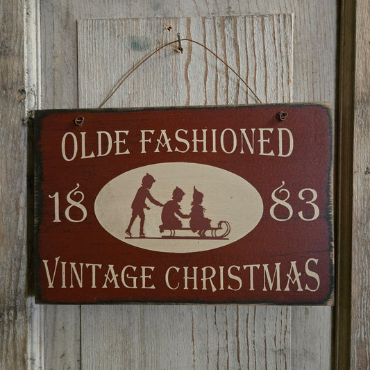 A rustic rectangular wooden hanging sign with a dark red painted background and cream lettering that reads “OLDE FASHIONED 1883 VINTAGE CHRISTMAS,” plus a cream oval with silhouetted figures in a sleigh. The sign hangs against a weathered wood plank wall.