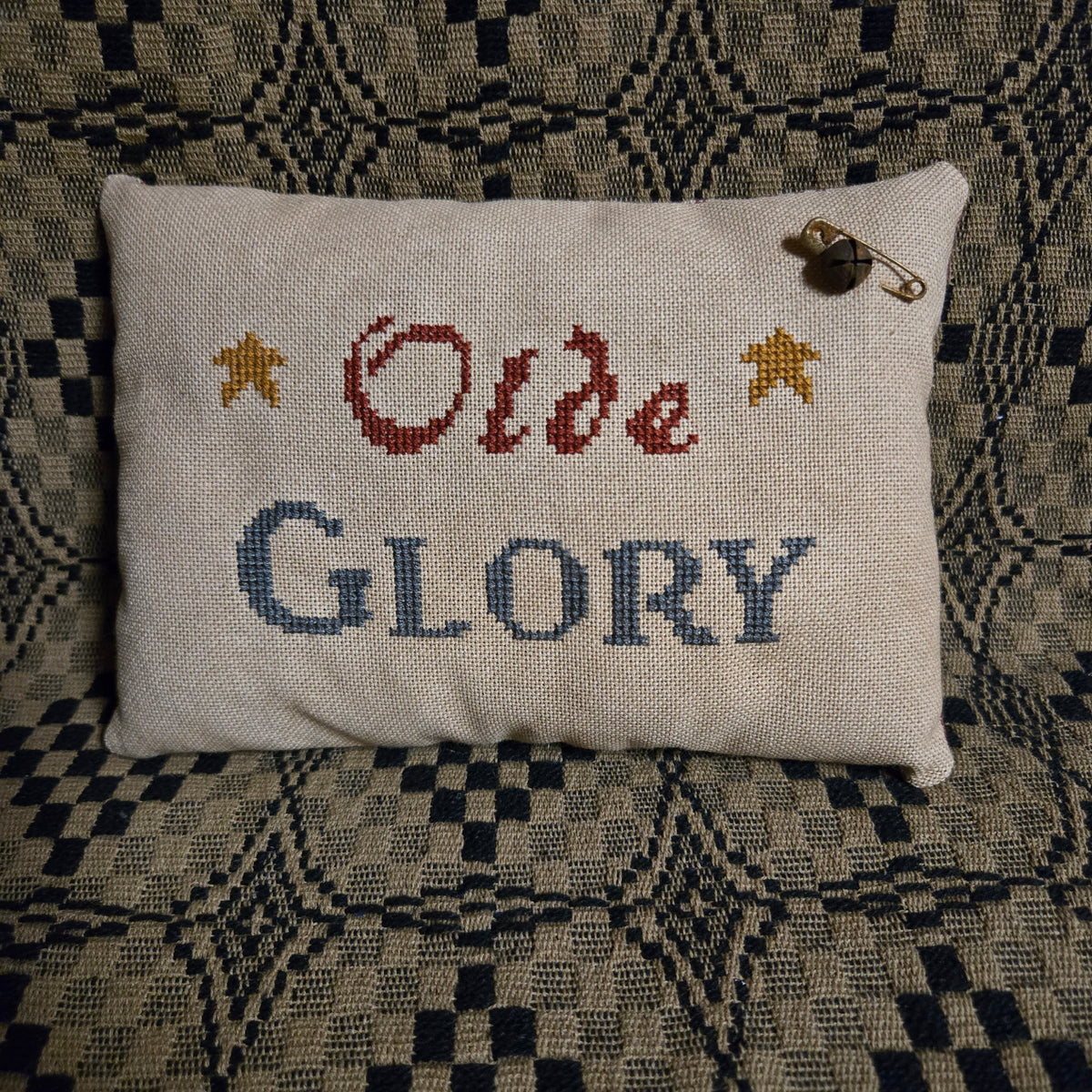 A square cream-colored cross-stitch pillow with the words 'Olde GLORY' stitched in red and blue, flanked by two gold stars. A small safety pin is attached near the top right corner.