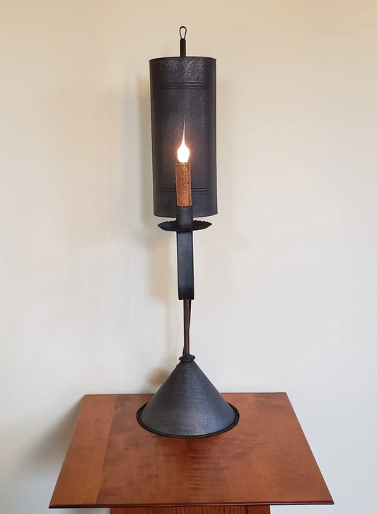 A black tin table light with a lit candle-like bulb, mounted on a wooden table against a plain wall.