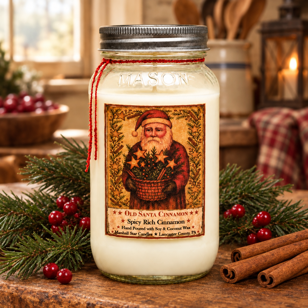 A glass jar candle with a metal lid and a red ribbon bow, featuring a vintage-style Santa illustration and the scent name 'Old Santa Cinnamon.' The candle is styled on a wooden surface with pine branches, cinnamon sticks, and red berries in a warm, rustic indoor setting.