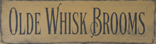 A rectangular wooden sign with the words 'OLDE WHISK BROOMS' in dark lettering, placed on a worn wooden surface.