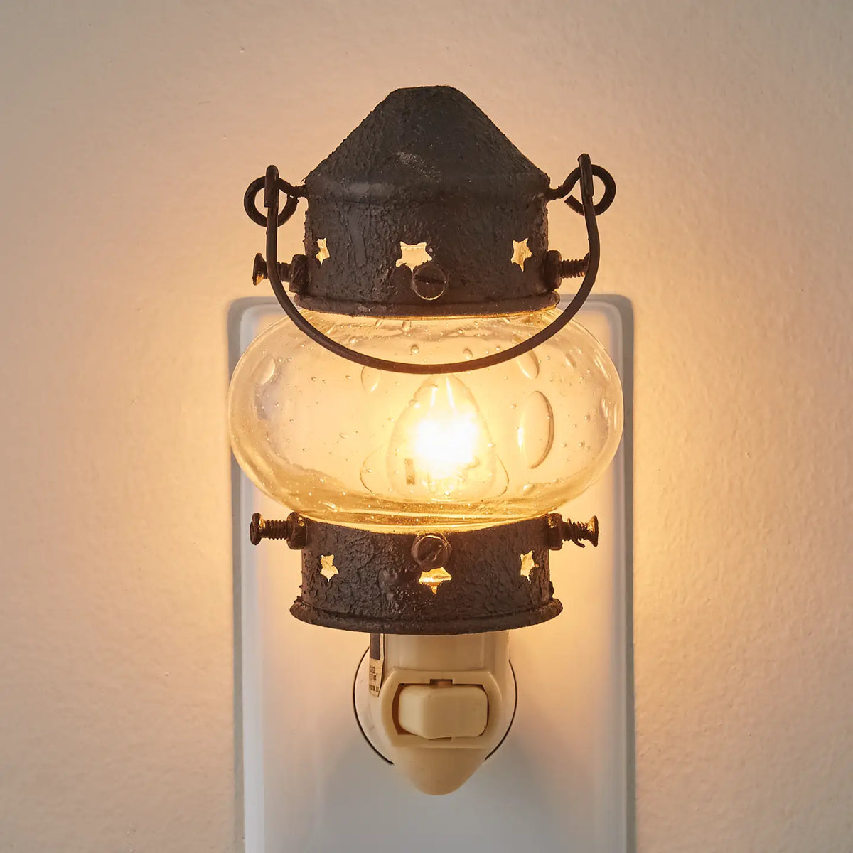 A vintage-style lantern night light with a glowing warm bulb inside a clear glass globe, mounted on a white wall switch. The lantern features a dark metal top and bottom with star-shaped cutouts and a curved handle.