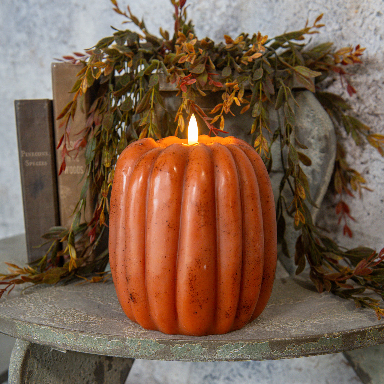 Pumpkin-shaped candle on a rustic surface with autumn leaves and books in the background