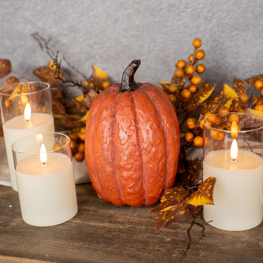 A tall, orange pumpkin-shaped decoration with a rustic texture, displayed in front of two lit candles, surrounded by autumn leaves and berries.