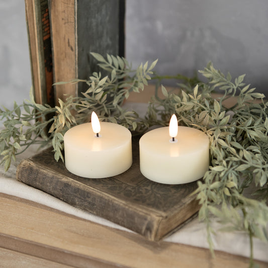 Two oversized 3D flame cream tealight candles on a wooden surface with a plant wreath behind them.