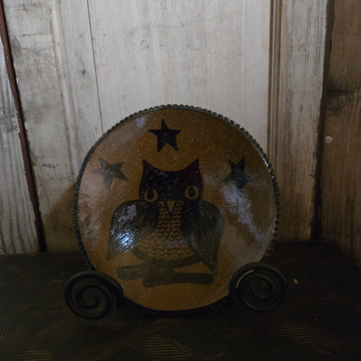 A rustic hand-painted ceramic plate featuring a black owl and stars on a brown speckled background, displayed on a black metal stand against a wooden paneled wall.