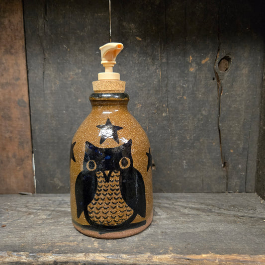 A rustic ceramic soap dispenser with a brown speckled body, a black and gold owl design, and a cork stopper with a cream-colored pump, set against a dark wooden background.
