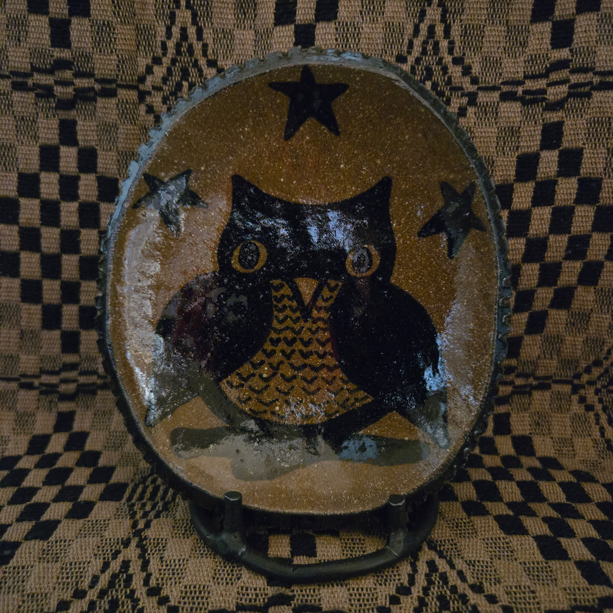 A round decorative ceramic plate featuring a hand-painted black owl with a patterned yellow chest, set against a brown background with black stars. The piece is displayed on a small stand in front of a black and tan checkered woven textile backdrop.