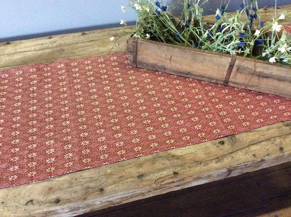 Red floral patterned table runner on a wooden table with a wooden planter box of flowers in the background.